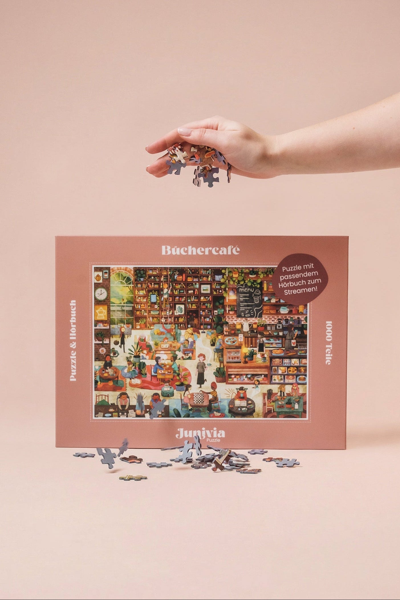 Junivia Büchercafé Puzzle: Hand lässt Puzzleteile über unserem Büchercafé Puzzle mit passendem Hörbuch fallen mit Buchladenmotiv und Selbstfindungsgeschichte 